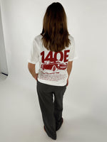 En person står med ryggen til iført en hvid T-shirt med rød grafik af en bil og tekst "140E Racing Club member 140E ... 1987 ... Car racing ...". De er i en lys indendørs omgivelser.