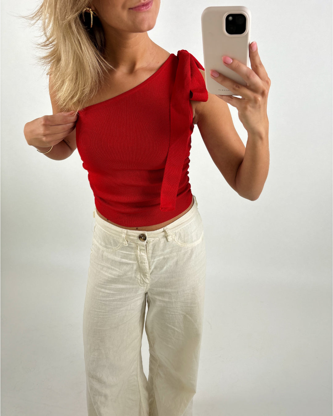 Kvinde i rød, ærmeløs top tager en selfie med telefon i højre hånd, iført hvide bukser, mod en neutral baggrund. Hun retter på sin top med venstre hånd.