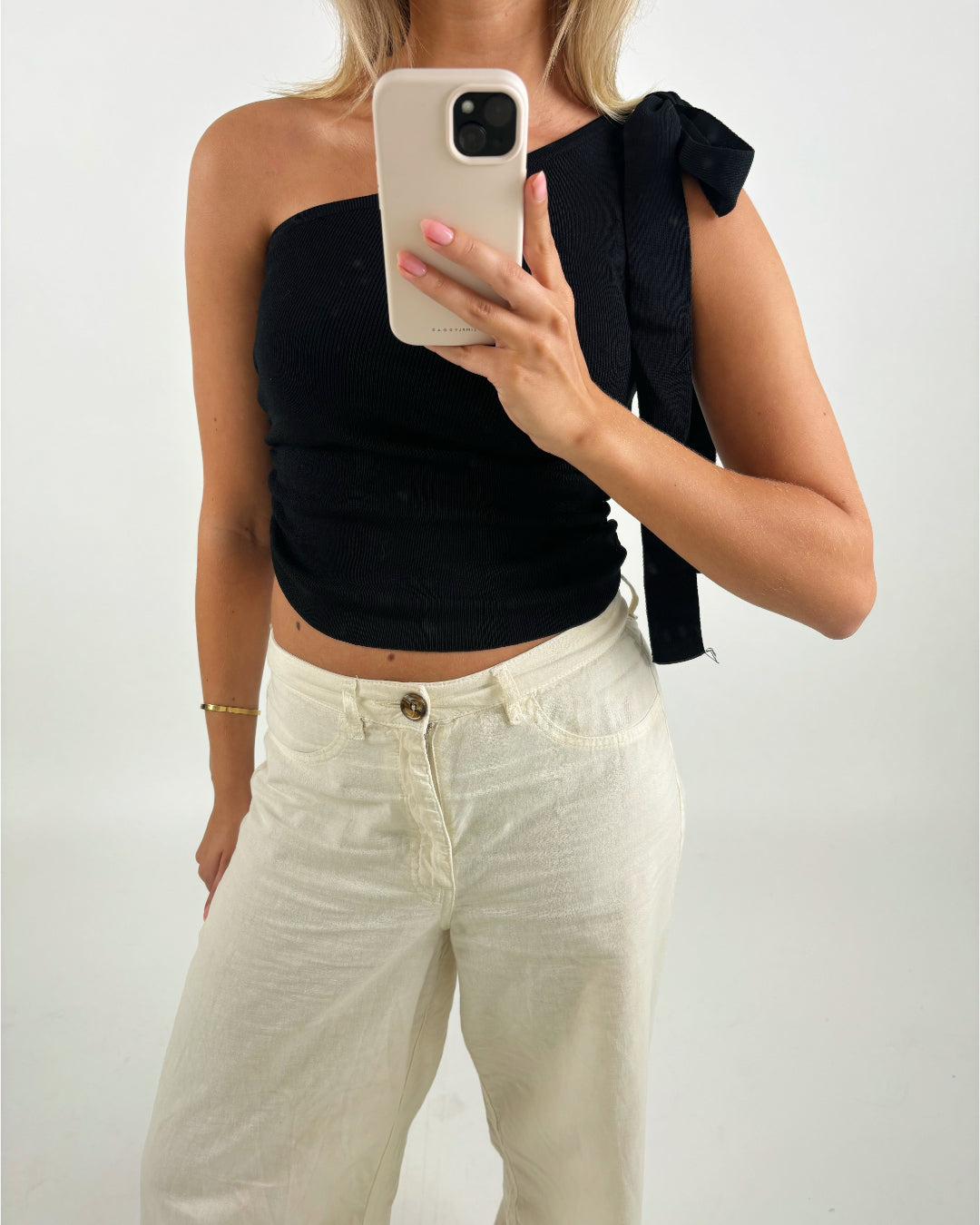 En person iført sort én-skulder top og hvide bukser tager et spejlbillede med en smartphone i hånden. Baggrunden er enkel og lys.