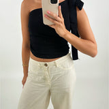 En person iført sort én-skulder top og hvide bukser tager et spejlbillede med en smartphone i hånden. Baggrunden er enkel og lys.