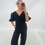 En person klædt i sort tager en selfie med en smartphone. Personen har håret sat op og bærer en halskæde, stående i et minimalistisk, lyst rum.