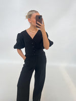 En person klædt i sort tager en selfie med en smartphone. Personen har håret sat op og bærer en halskæde, stående i et minimalistisk, lyst rum.