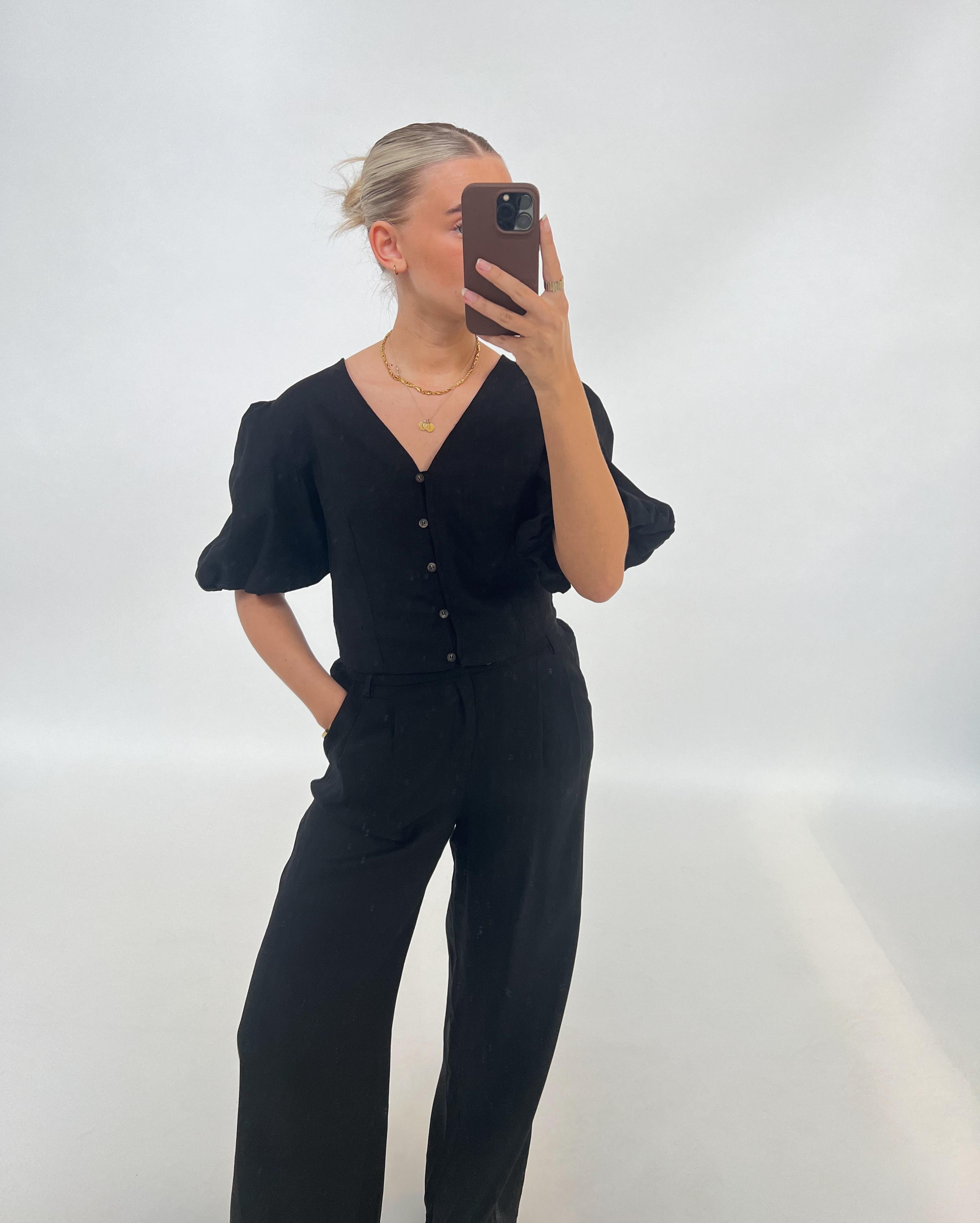 En person klædt i sort tager en selfie med en smartphone. Personen har håret sat op og bærer en halskæde, stående i et minimalistisk, lyst rum.