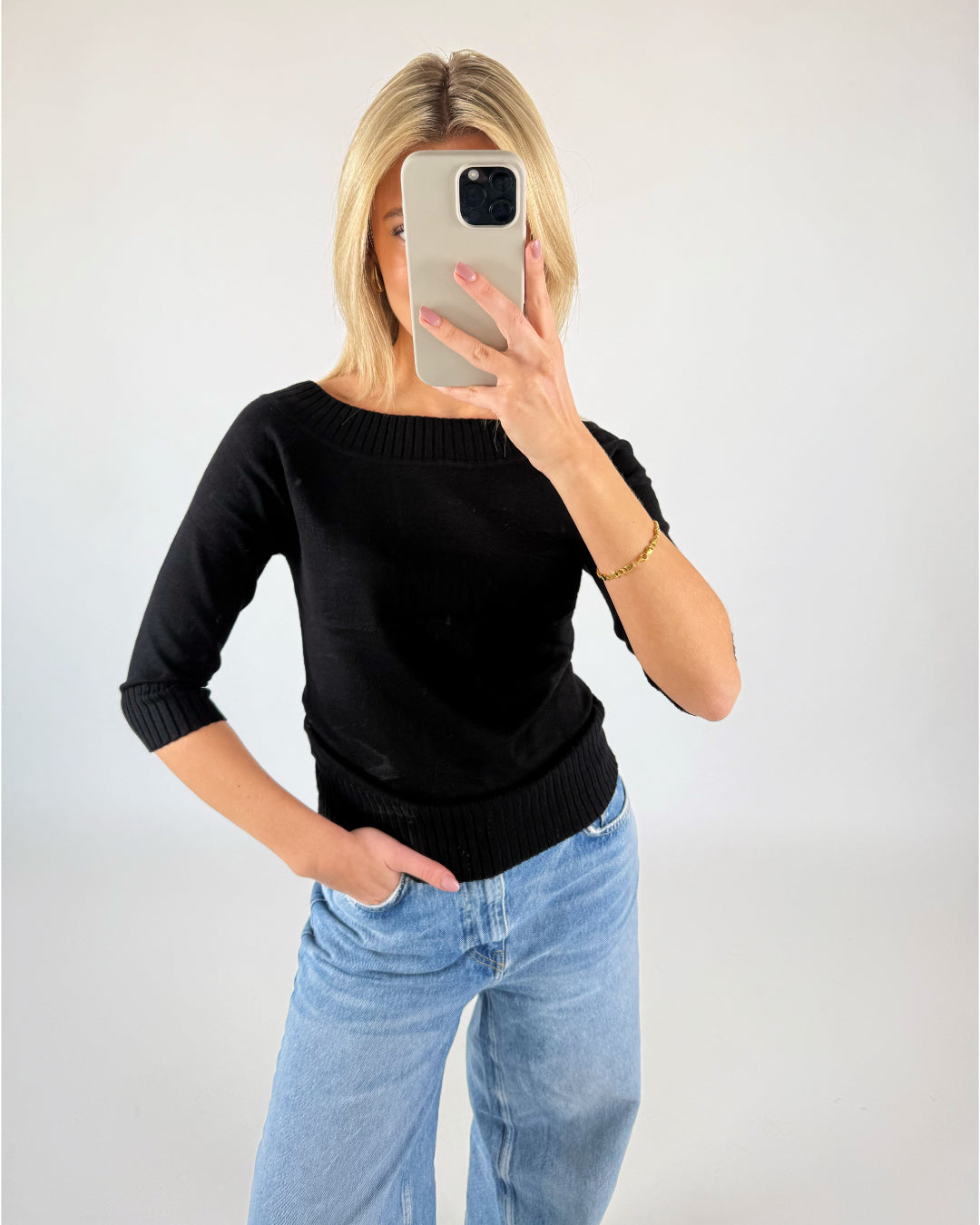 Person står foran et kamera med spejl, klædt i sort bluse og lyseblå jeans, tager et selfie. Lyse vægge omkranser personen i et neutralt miljø.