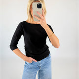 Person står foran et kamera med spejl, klædt i sort bluse og lyseblå jeans, tager et selfie. Lyse vægge omkranser personen i et neutralt miljø.