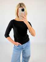 Person står foran et kamera med spejl, klædt i sort bluse og lyseblå jeans, tager et selfie. Lyse vægge omkranser personen i et neutralt miljø.