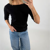 En kvinde iført sort trøje og lyse jeans tager et spejlbillede med sin smartphone i et lyst rum. Hun har en hånd i lommen og bærer et armbånd.
