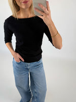 En kvinde iført sort trøje og lyse jeans tager et spejlbillede med sin smartphone i et lyst rum. Hun har en hånd i lommen og bærer et armbånd.