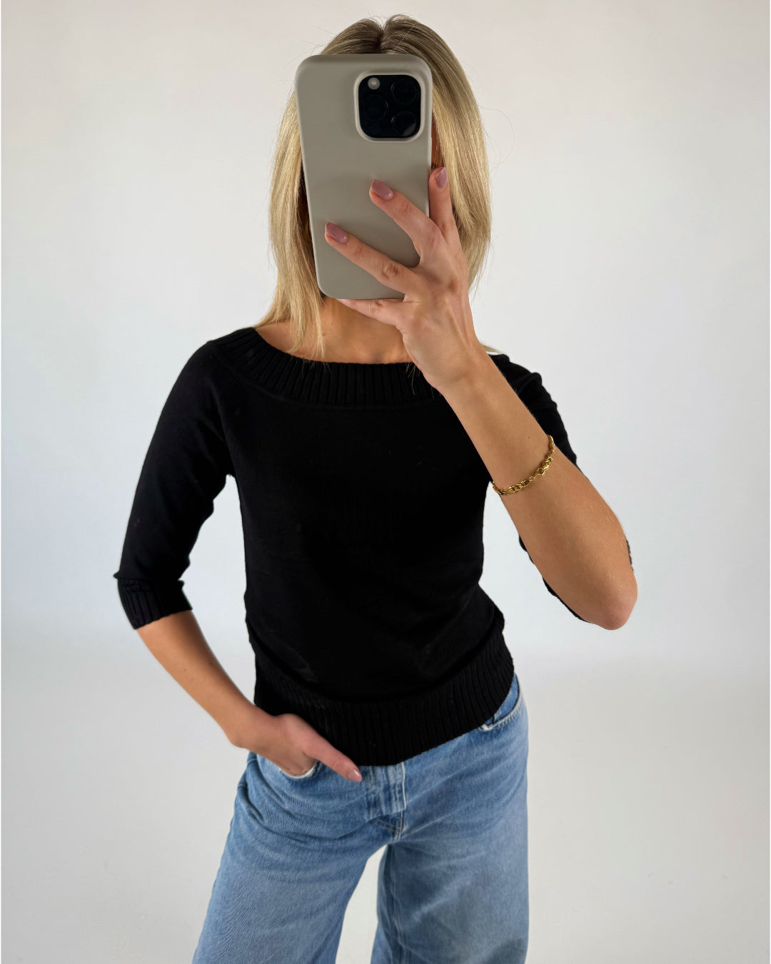 En person iført sort trøje og blå jeans tager en selfie med en mobiltelefon i et minimalistisk, hvidt rum.