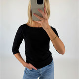 En person iført sort trøje og blå jeans tager en selfie med en mobiltelefon i et minimalistisk, hvidt rum.