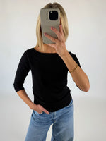 En person iført sort trøje og blå jeans tager en selfie med en mobiltelefon i et minimalistisk, hvidt rum.