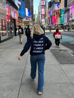 En person med en "Los Angeles"-trøje går på en travl byfortov om dagen. Omkring ses andre fodgængere, skyskrabere og store reklameskærme.