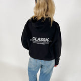 En person bærer en sort hættetrøje med teksten "CLASSIC" i hvid. Personen står med ryggen til, iført lyseblå jeans, i et hvidt rum.