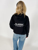 En person bærer en sort hættetrøje med teksten "CLASSIC" i hvid. Personen står med ryggen til, iført lyseblå jeans, i et hvidt rum.