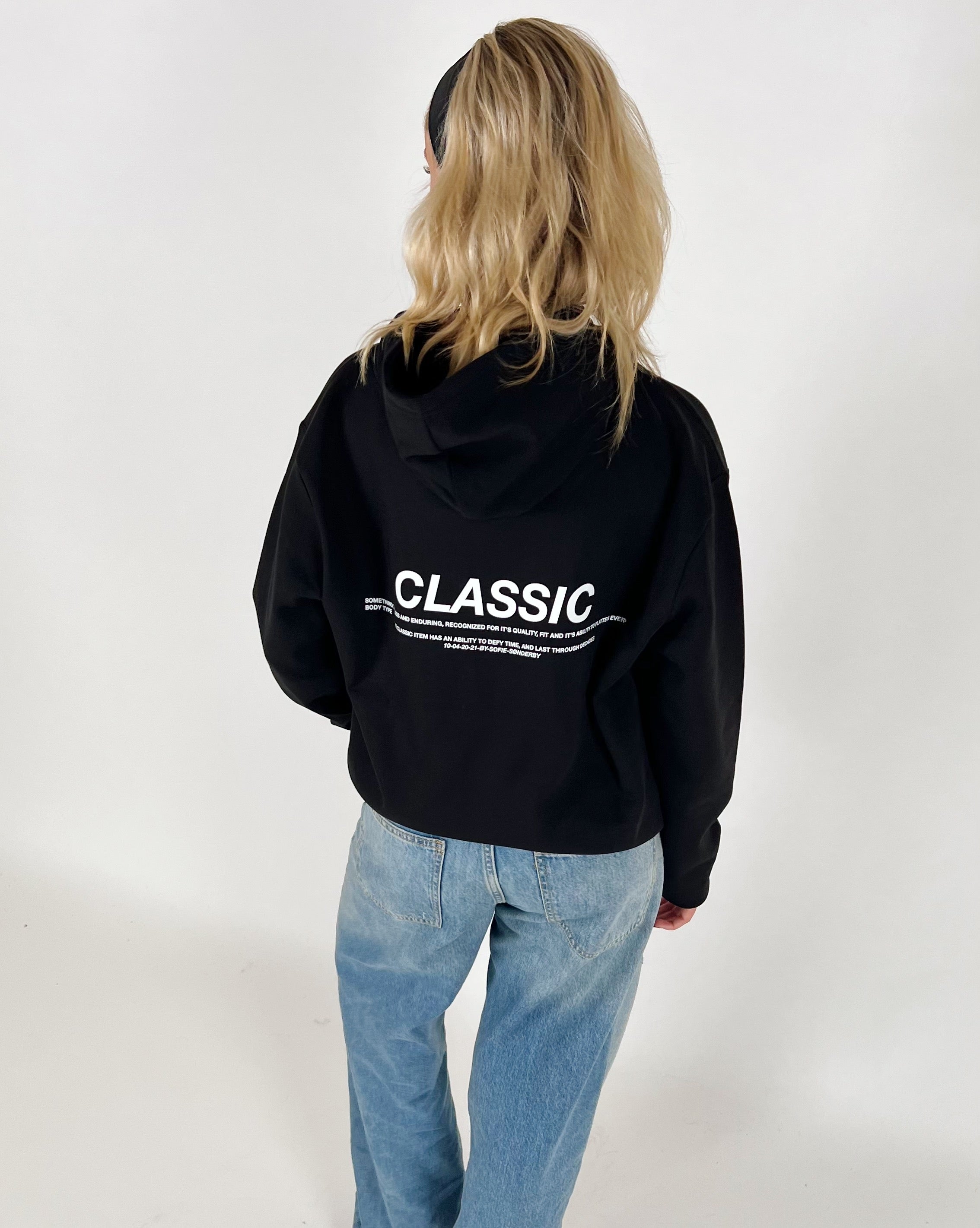 En person bærer en sort hættetrøje med teksten "CLASSIC" i hvid. Personen står med ryggen til, iført lyseblå jeans, i et hvidt rum.