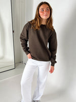 Kvinde iført brun sweatshirt og hvide bukser står afslappet i et lyst værelse, med en spejlvendt dør i baggrunden. Hun kigger let til siden.