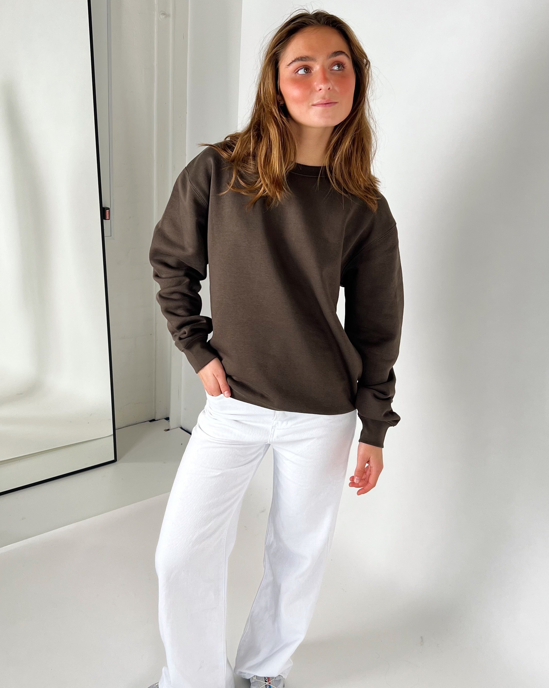 Kvinde iført brun sweatshirt og hvide bukser står afslappet i et lyst værelse, med en spejlvendt dør i baggrunden. Hun kigger let til siden.