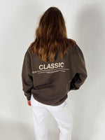 En person iført en brun sweatshirt med teksten "CLASSIC" og yderligere lille skrift. Personen står med ryggen til, klædt i hvide bukser, mod en hvid baggrund.