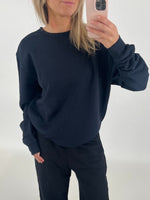 En person iført sort sweatshirt tager et spejlfoto med en smartphone i en lys, minimalistisk baggrund.