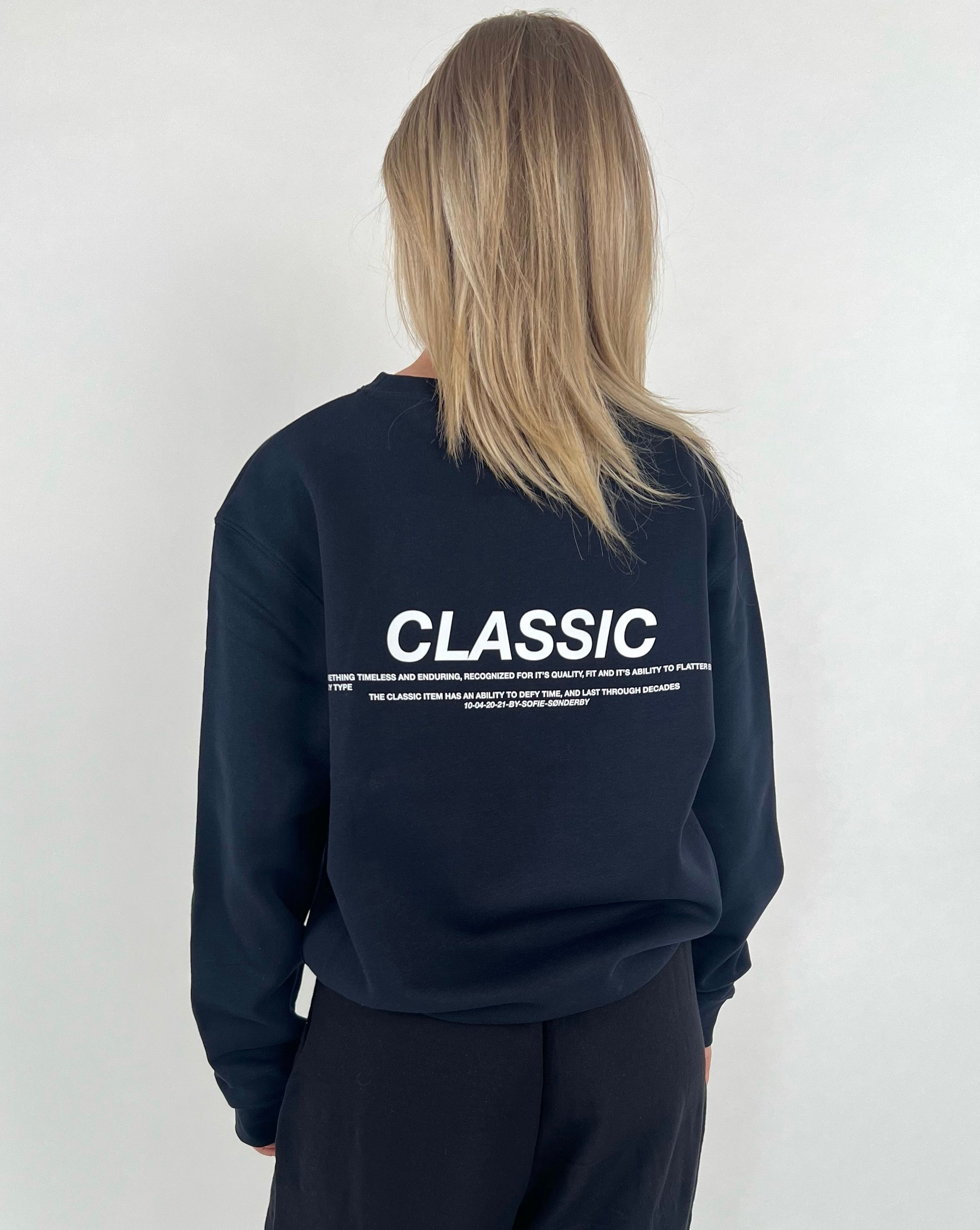 En person står med ryggen til iført en sort sweatshirt med teksten "CLASSIC". Omgivelserne er enkle og neutrale med en lys baggrund.