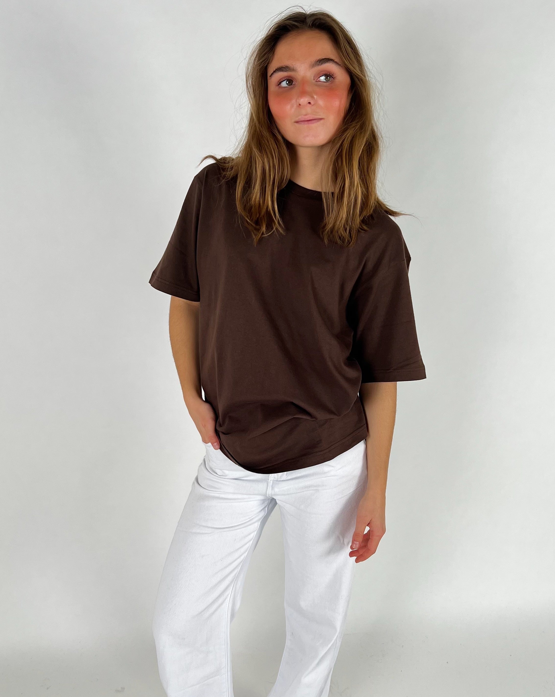 En person iført brun t-shirt står med én hånd i lommen på hvide bukser mod en hvid baggrund. Personen kigger til siden, og håret er løst.