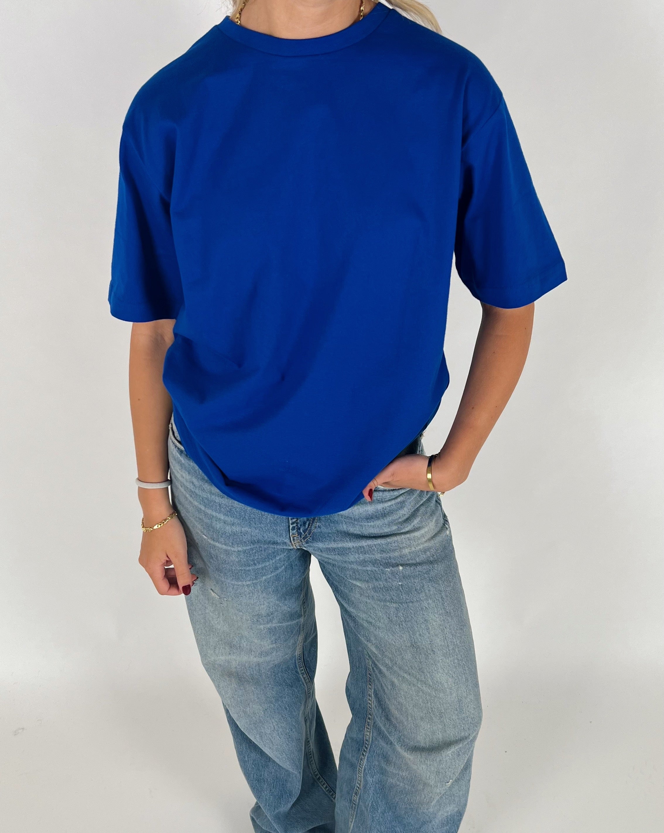 Person iført en blå T-shirt og jeans står med hånden i lommen. De har armbånd og rød neglelak, i en lys, minimalistisk baggrund.