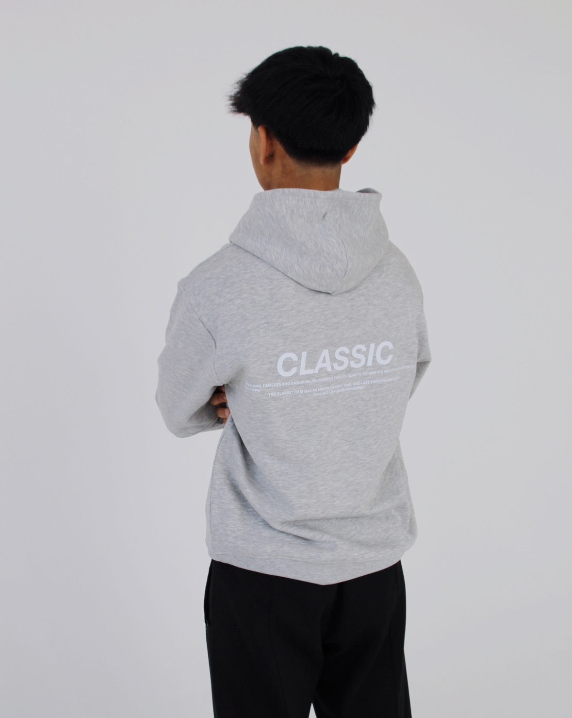 En person iført en grå hættetrøje med teksten "CLASSIC" står med krydsede arme i et hvidt rum.