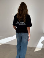 En person står med ryggen til iført en sort t-shirt med teksten "CLASSIC" og blå jeans i et oplyst rum uden dekoration.