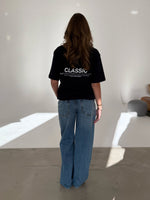En person med langt hår og blå jeans står stille med ryggen til, iført en sort T-shirt med teksten "CLASSIC" i et minimalistisk, lyst rum.