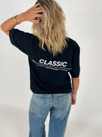 Kvinde i sort T-shirt med teksten "CLASSIC" står med ryggen til, rører sit hår. Hun bærer lyse jeans i en neutral, lys baggrund. Teksten på T-shirten er delvist læsbar.
