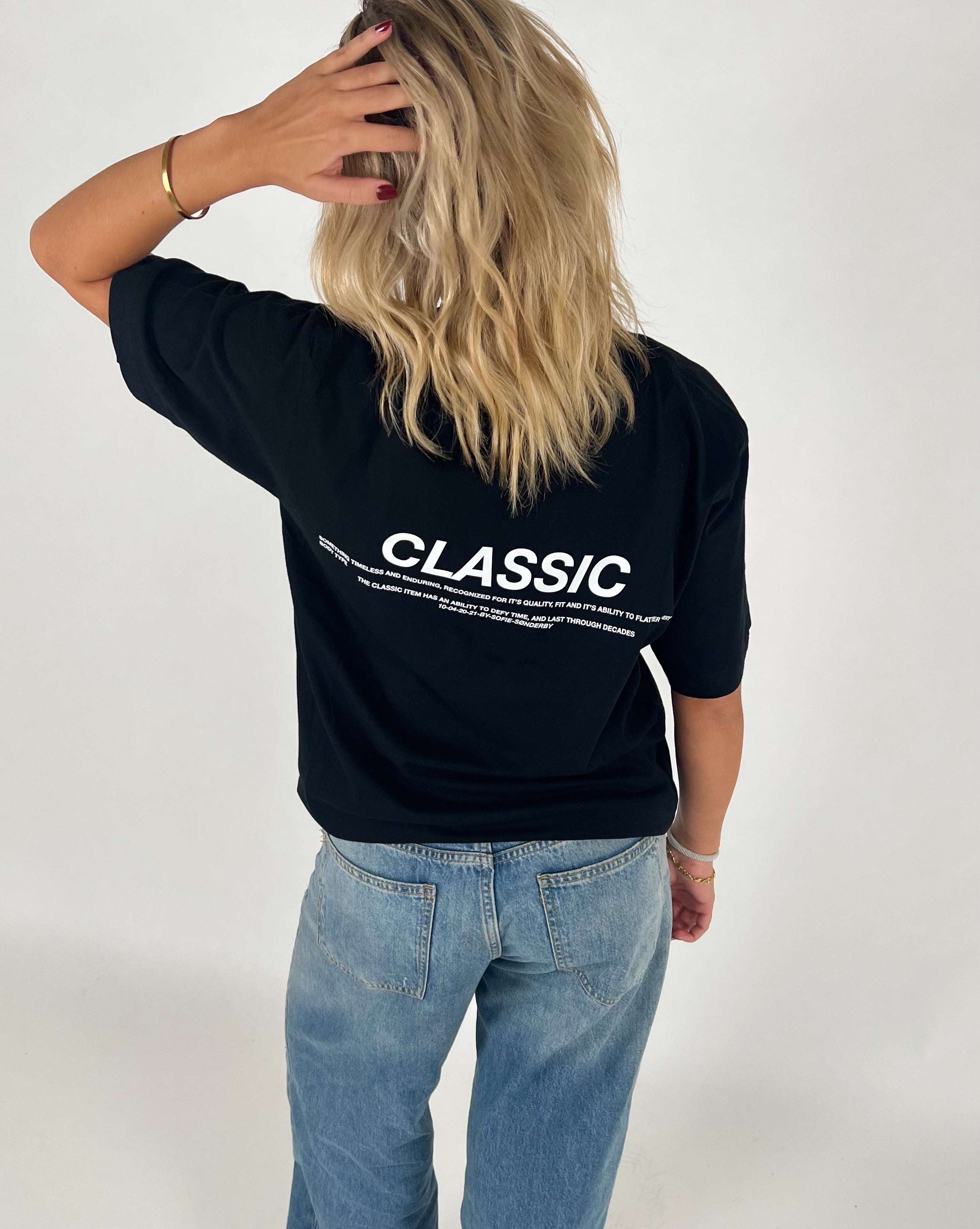 Kvinde i sort T-shirt med teksten "CLASSIC" står med ryggen til, rører sit hår. Hun bærer lyse jeans i en neutral, lys baggrund. Teksten på T-shirten er delvist læsbar.