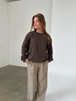 Kvinde iført brun sweatshirt og beige bukser står afslappet med hænderne i lommerne i et rum med hvide murstensvægge og gråt gulv.