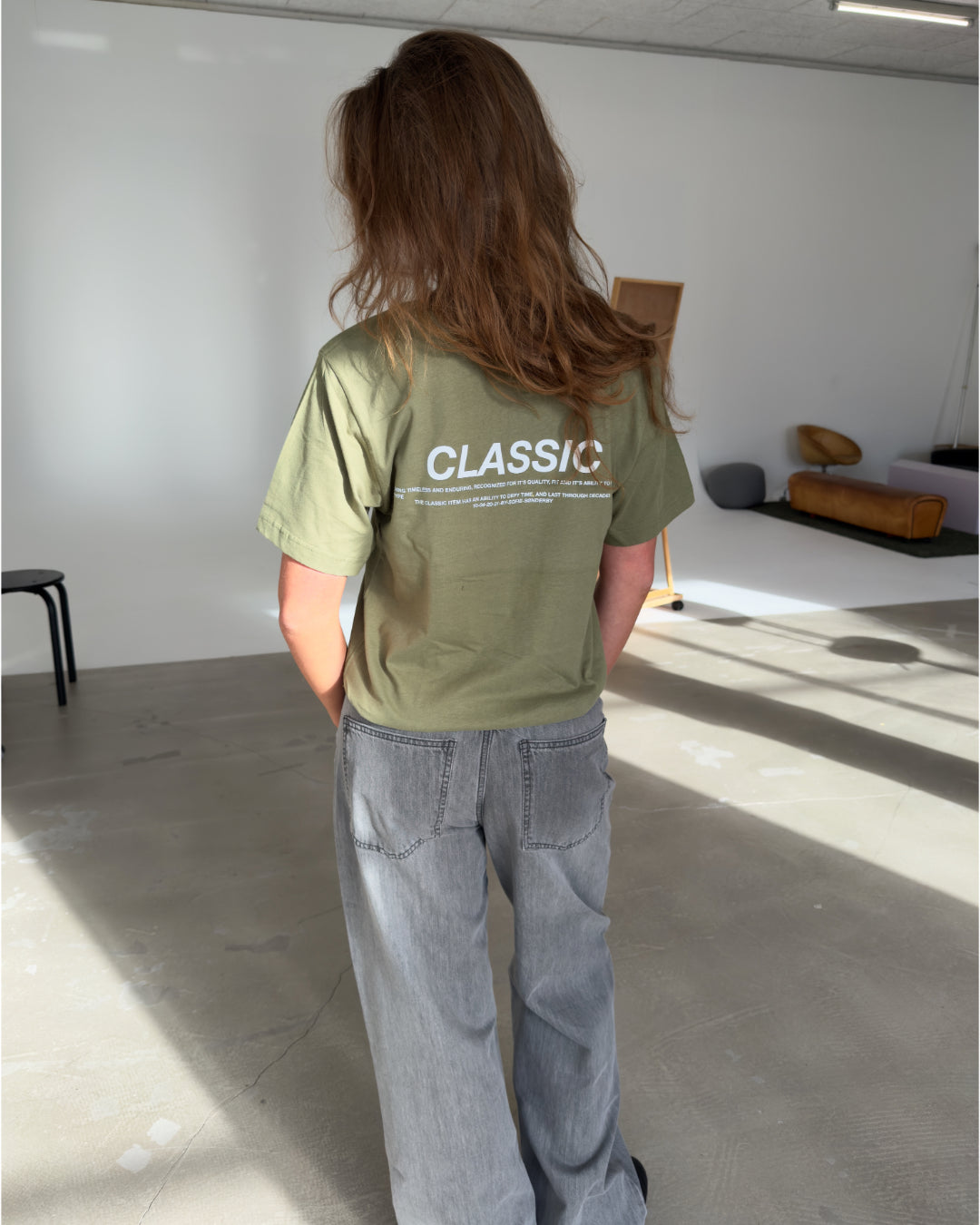 En person med langt brunt hår står i et lyst rum iført en grøn t-shirt med teksten "CLASSIC" på ryggen og grå jeans. Rummet har minimalistisk møblering.
