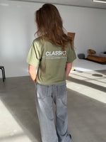 En person med langt brunt hår står i et lyst rum iført en grøn t-shirt med teksten "CLASSIC" på ryggen og grå jeans. Rummet har minimalistisk møblering.