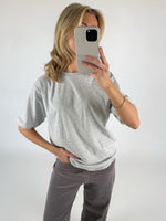 En person iført grå T-shirt og mørke bukser holder en smartphone foran ansigtet. Personen står i et lyst rum og ser ud til at tage et spejlbillede.