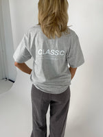 En person iført en grå T-shirt og mørke bukser står med ryggen til. På T-shirten står teksten "CLASSIC". Baggrunden er lys og enkel.