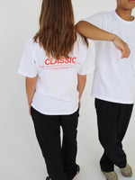 To personer iført hvide T-shirts og sorte bukser står mod en lys baggrund. Den ene T-shirt har tekst på ryggen: "CLASSIC" efterfulgt af yderligere ord i mindre skrift.