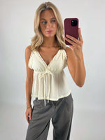 En person tager en selfie iført en lyseblå top og grå bukser. Personen står i et rum med en lys baggrund og holder en rød mobiltelefon.