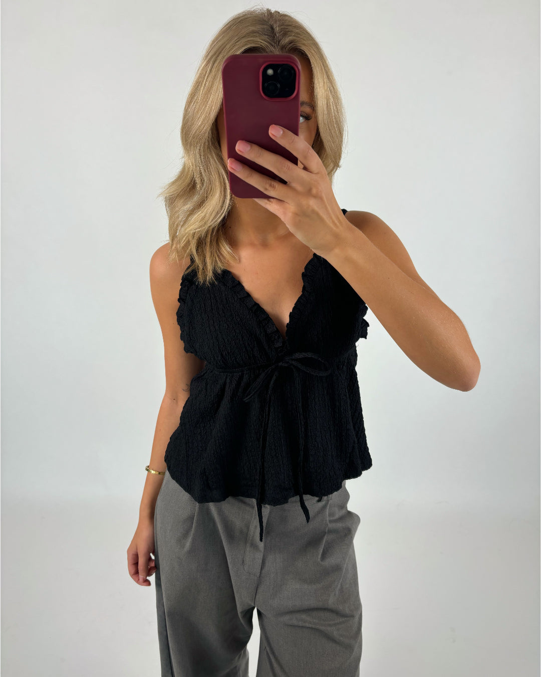 En person tager en selfie med en mobiltelefon iført en sort top og grå bukser. Baggrunden er hvid, hvilket fremhæver tøjet og skaber en minimalistisk kontekst.