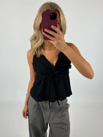 En person tager en selfie med en mobiltelefon iført en sort top og grå bukser. Baggrunden er hvid, hvilket fremhæver tøjet og skaber en minimalistisk kontekst.