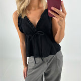 En person iført en sort bluse med flæsedetaljer og et bånd ses tage en selfie. Personen har blondt hår og bærer grå bukser. Baggrunden er neutral.