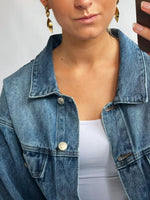 En person bærer en denimjakke og store gyldne øreringe. De tager et selfie i spejlet med pastelfarvet baggrund. Jakken er åben over en hvid top.