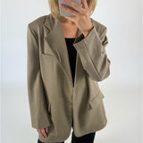 En person iført en beige blazer holder en smartphone op foran ansigtet. Personen står i et rum med en neutral, hvid baggrund.