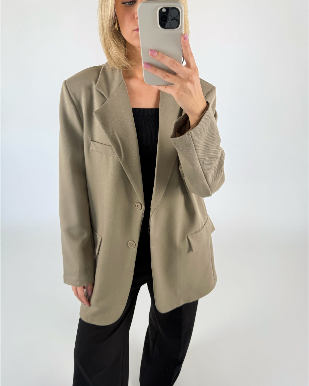 En person i beige blazer tager et spejlbillede med en smartphone, iført sort top og bukser; omgivelserne er et hvidt, enkelt rum.