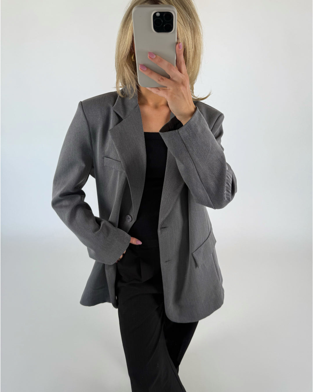 En person i gråt jakkesæt tager et spejlfoto med en smartphone i et neutralt oplyst rum. Personen har blondt hår og sort tøj under jakken.