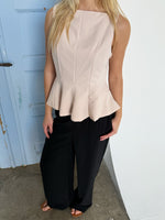 En kvinde står iført en lys top med peplum og sorte bukser, med en blå dør i baggrunden.