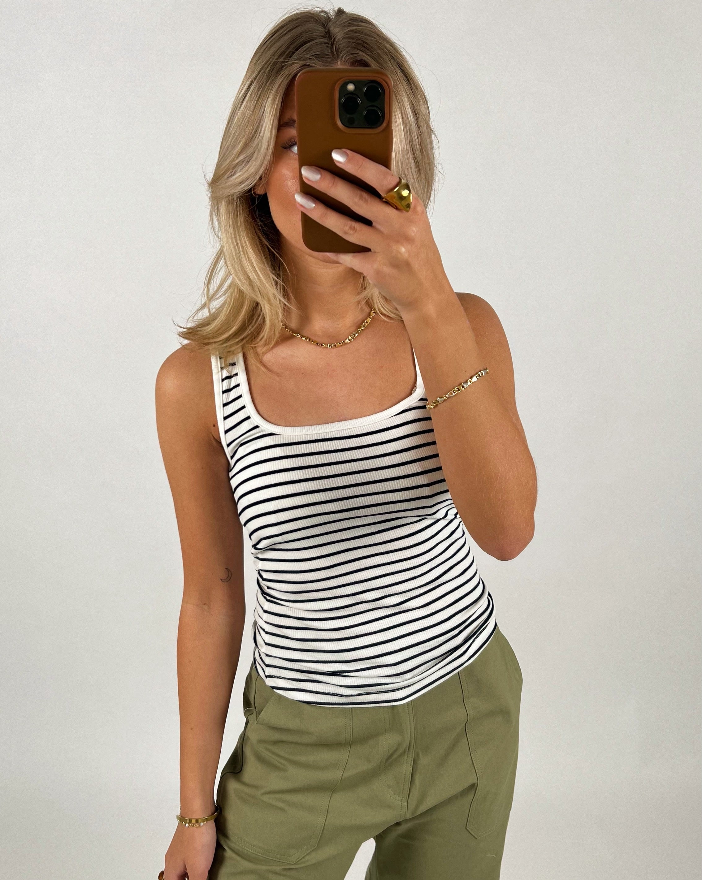 En person tager en selfie med en mobiltelefon, iført en sort/hvid-stribet tanktop og grønne bukser, mod en neutral baggrund.