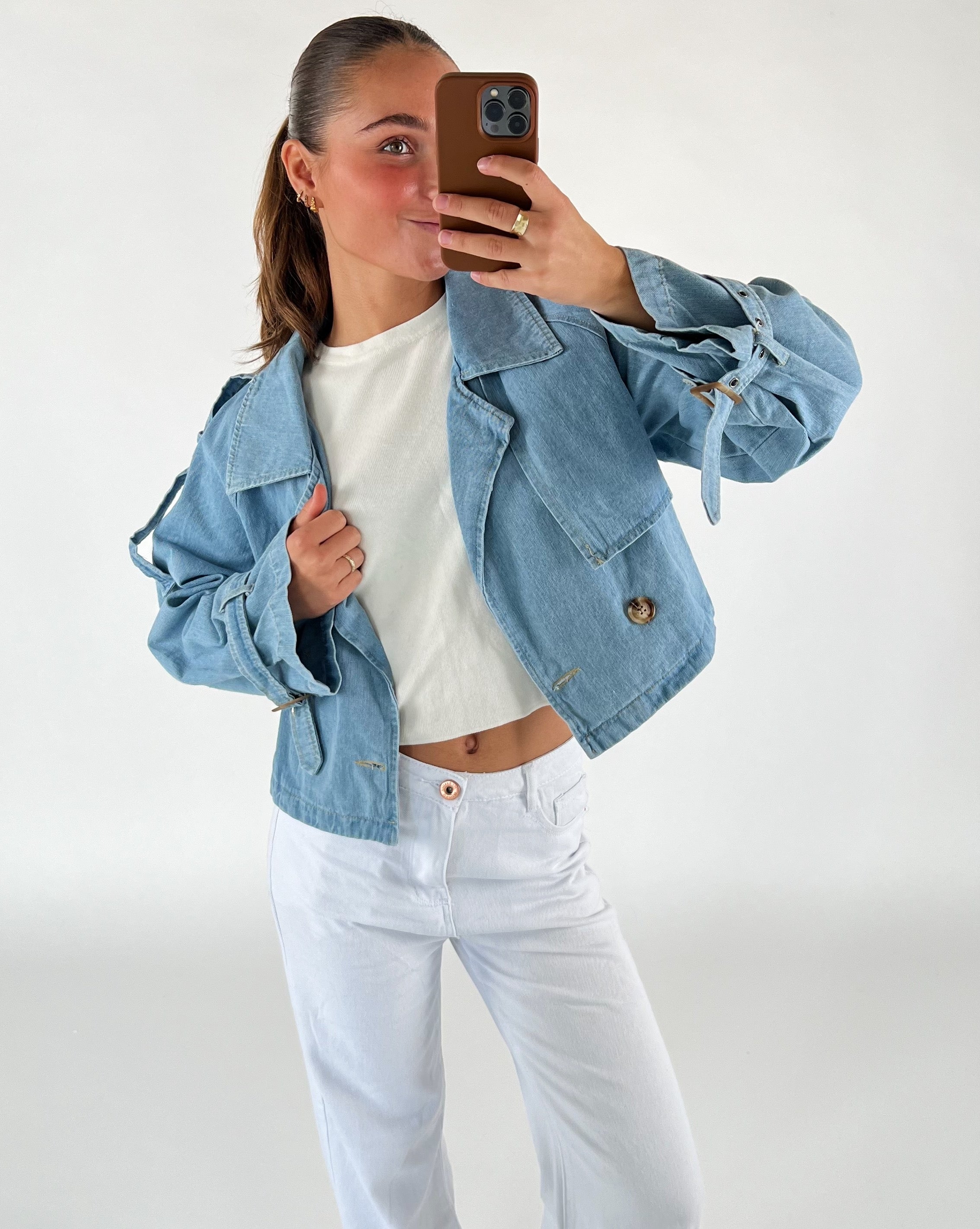 En person står foran et spejl, iført en lyseblå jakke og hvide bukser, tager en selfie med en brun mobiltelefon i en minimalistisk indendørs setting uden synlig tekst.