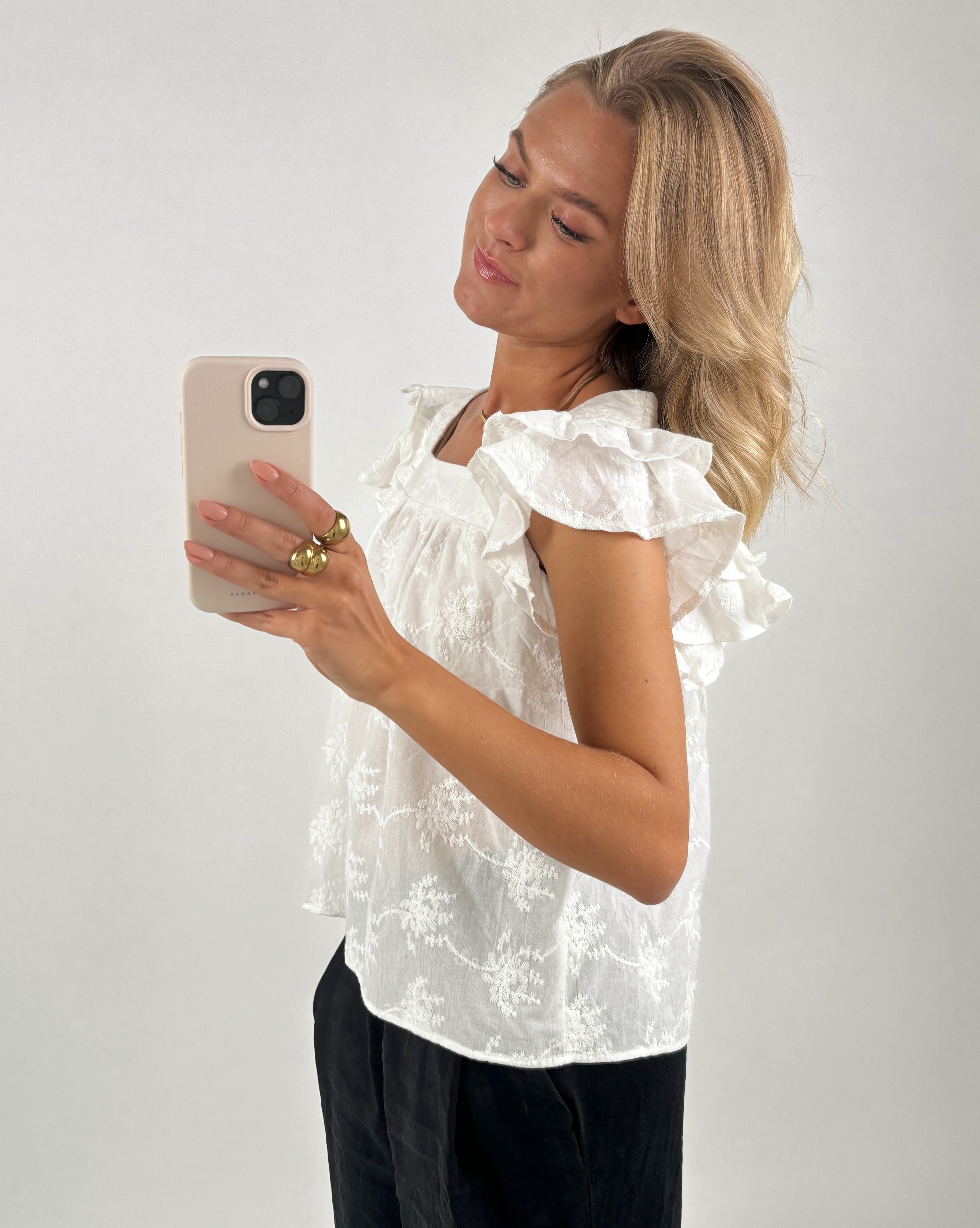 En kvinde i en hvid, mønstret bluse tager et spejlbillede med sin smartphone. Hun står i en minimalistisk indrettet lys baggrund. Hun bærer guldringe.