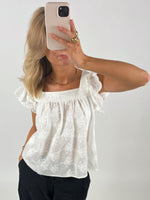 En person iført hvid, mønstret bluse tager et spejlselfie med en smartphone. Personen har let bølget, blond hår og guldtilbehør. Baggrunden er en ensfarvet væg.